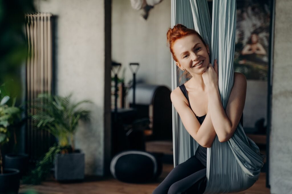 Happy red haired woman sitting in comfy position in yoga hammock